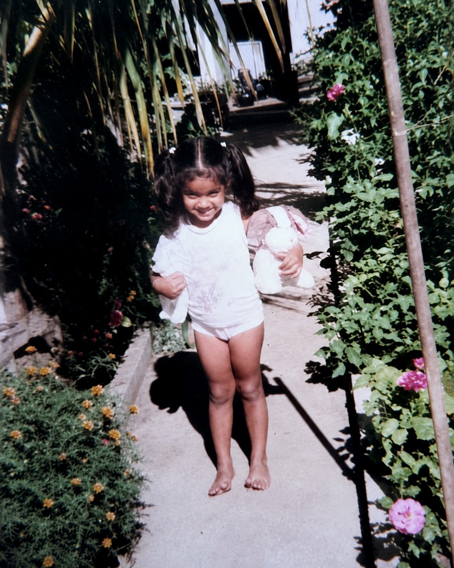 Karoona as a child, walking barefoot along a garden path, holding a soft toy.